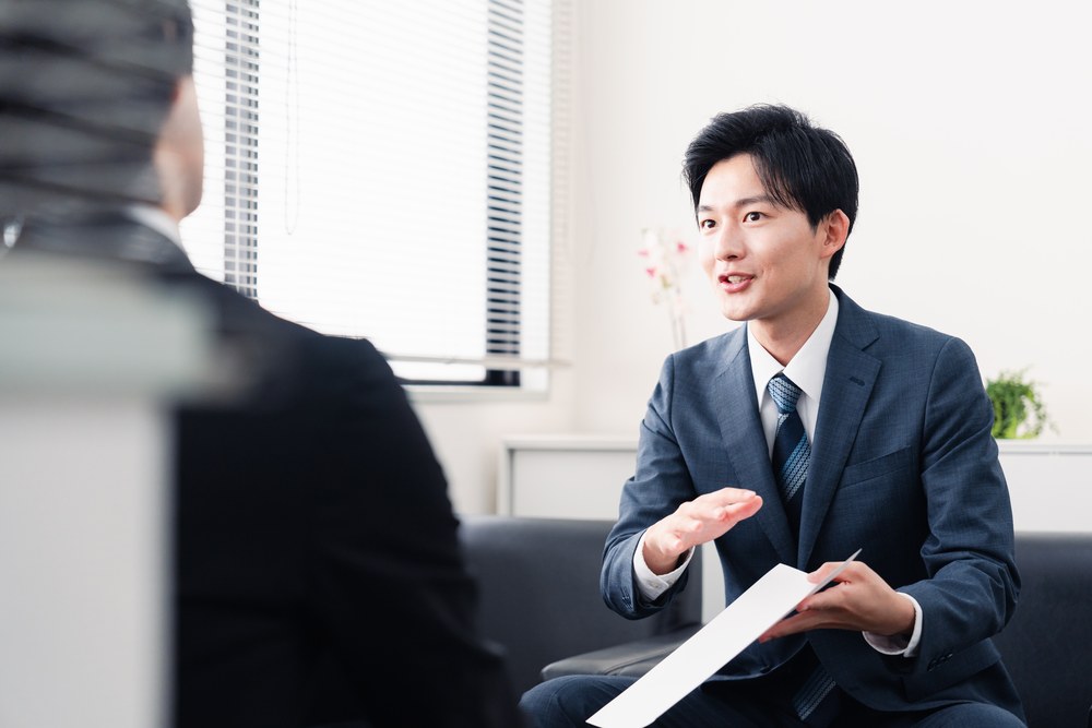 小さな事務所のソファで、書類を示しながら説明する日本人の担当者。ファクタリング会社選びの比較ポイントを解説する記事冒頭の写真
