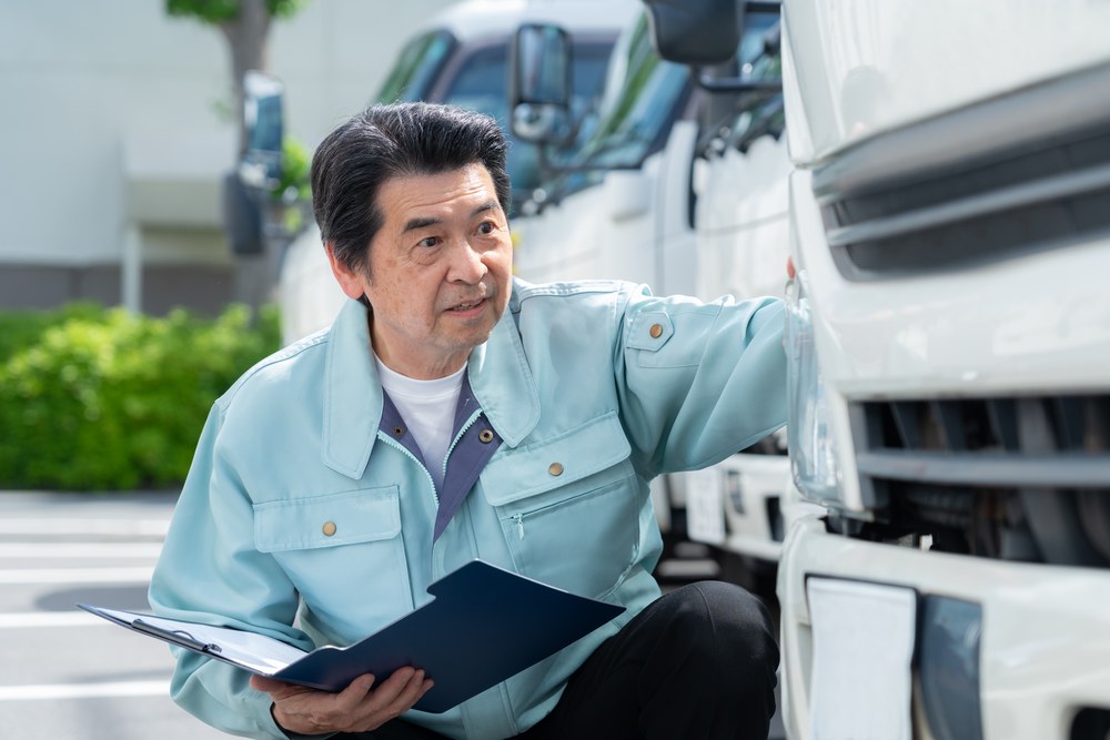 運送会社の車庫でトラックの点検をする作業着姿の日本人ドライバー。運賃の入金待ちが長い悩みを解説する記事冒頭の写真
